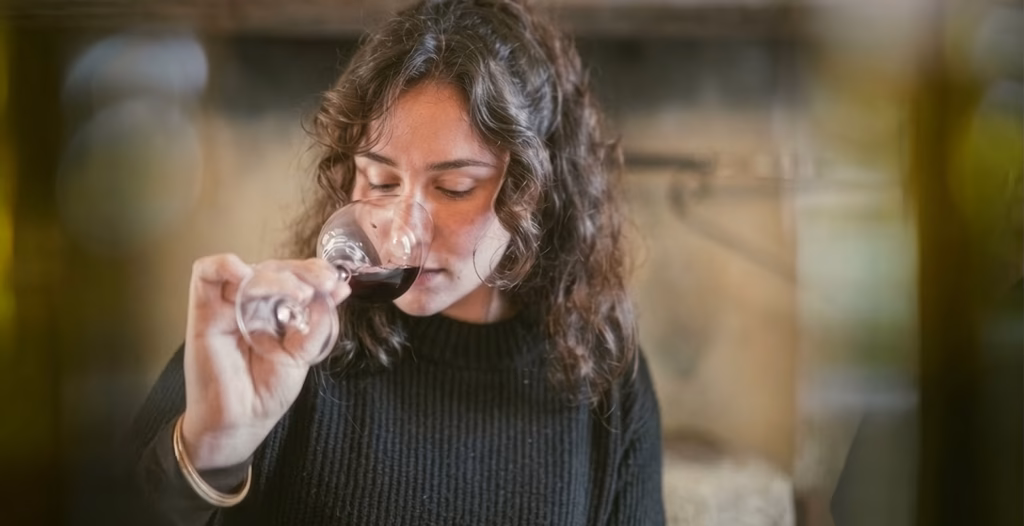 Woman with curly hair in a black sweater smelling red wine in a glass.