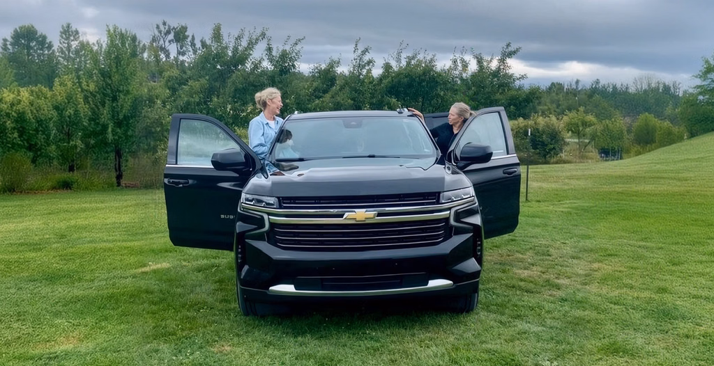 Two women standing beside a black Chevrolet SUV with open doors on a grassy field.