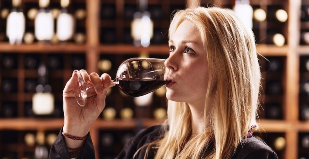 A blonde woman sipping red wine from a glass in a wine cellar with wooden bottle racks in the background.