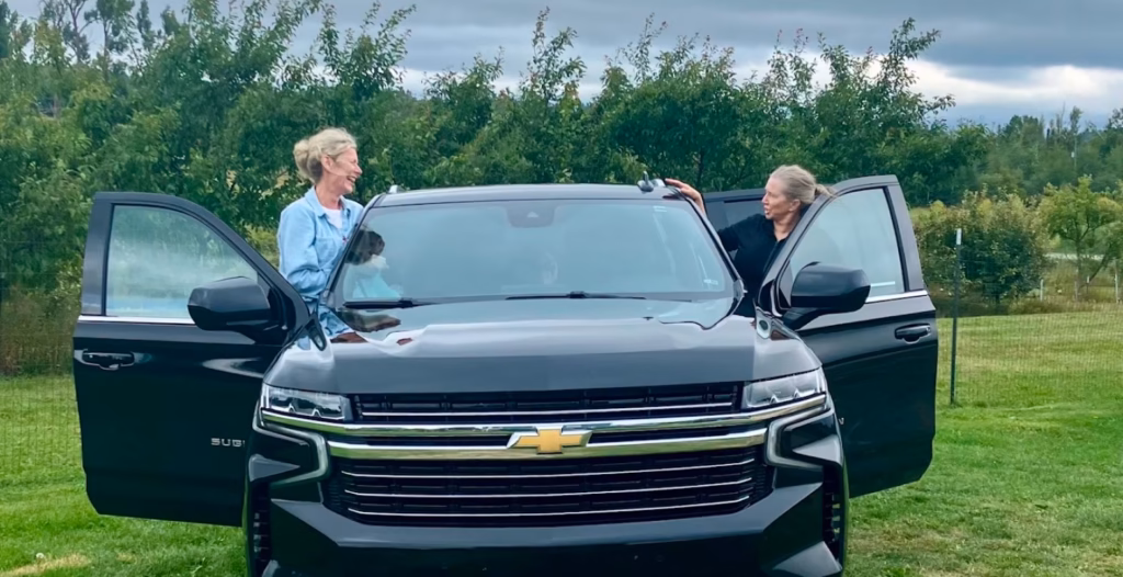 Two women chatting while getting out of a black Chevrolet Suburban parked on a grassy field surrounded by trees.